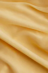 Camisa de malla color amarillo pálido, de corte entallado, con cuello clásico, mangas largas con puños abotonados y cierre frontal con botones a presión ocultos. Presenta frunces en la parte delantera que crean un efecto drapeado.