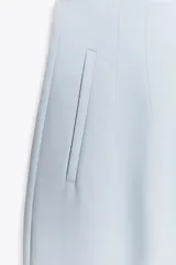 Pantalón de vestir color crema, de tiro alto, corte recto y largo cropped. Presenta bolsillos laterales de ojal y cierre frontal con cremallera, botón interior y gancho metálico.