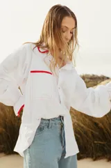 Camisa blanca de corte holgado con cuello camisero y mangas largas con puños elásticos. Presenta bolsillo de parche en el pecho y detalles de ribete rojo en cuello, puños y bolsillo.
