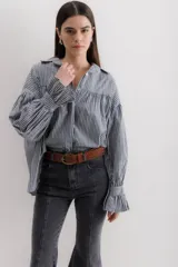 Camisa gris a rayas verticales blancas, de corte amplio, cuello alto con lazo y mangas largas abullonadas con puños elásticos.