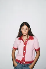 Chaqueta color blanco de punto con cuello solapa y manga corta. Tiene cierre frontal con botones color naranja y ribete en azul marino y naranja.