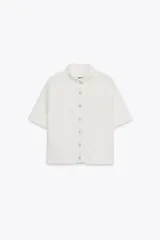 Chaqueta color blanco de tejido de punto, con cuello subido, manga corta y cierre frontal con botones.