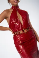 Vestido midi rojo de cuero con cuello halter, abertura en el pecho, cinturón de aros dorados y cierre lateral con cremallera.