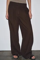 Pantalón marrón de tiro medio, corte ancho y largo cropped. Confeccionado en algodón, con cintura con trabillas y elástico en la espalda, bolsillos laterales y traseros, pinzas en el delantero y cierre frontal con cremallera, botón y ganchos metálicos.