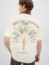 Remera color hueso de algodón con cuello redondo y manga corta. Estampado en la espalda con diseño de hojas de palmera y texto "Brezza Del Mattino Originals Summer Edition EST 1990".