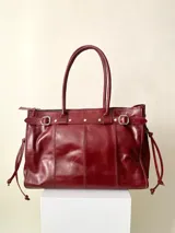 Bolso grande de cuero color vino tinto, con estructura rígida y asas para llevar al hombro. Presenta una tira decorativa con tachas y hebillas plateadas en la parte superior frontal, y lazos ajustables en los laterales. El interior no tiene forro y cuenta con un bolsillo grande tipo sobre con cierre.