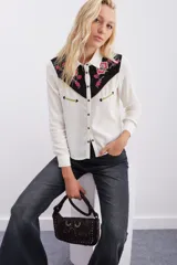 Camisa blanca de manga larga con canesú delantero y trasero en tela negra con bordados florales en tonos rosados y detalles en hilo dorado. Cierre frontal con botones metálicos tipo snap.