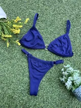 Bikini azul oscuro con tela texturada. El corpiño es triangular con una argolla de zamak en el centro y tirantes finos. La bombacha es colaless con argollas metálicas en los laterales.