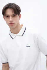 Chomba blanca de algodón con cuello clásico, media cartera de botones, bordado con la palabra 'Uniform' en el pecho y puños con rayas azul marino y rojo.