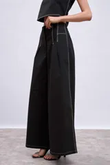 Pantalón negro de tiro alto, corte ancho y largo, con pliegues frontales y pespuntes contrastantes en blanco en la cintura y bolsillos.