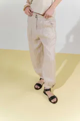 Pantalón de lino color beige con rayas verticales finas, corte barrel, tiro medio, pretina ancha con doble abotonadura, bolsillos laterales, pinzas en el frente y ruedo con presilla.
