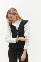 Chaleco blanco con finas rayas verticales negras, escote en V, cierre frontal con botones negros y volados en lino negro en los hombros. La parte inferior presenta otro volado en la misma tela del cuerpo.