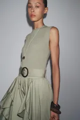 Vestido midi color verde oliva, de cuello redondo y sin mangas. Presenta un cinturón con hebilla metálica y detalle de volantes en la falda.