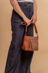 Bandolera de simil cuero color marrón topo, con textura ligeramente desgastada y detalles en nobuck. Presenta múltiples bolsillos frontales con cierres metálicos y tiradores de cuero. Correa ajustable para llevar al hombro o cruzada.