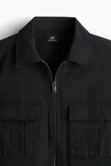 Sobrecamisa negra de algodón con cuello inglés, cierre bidireccional, bolsillos superiores con solapa y botones a presión, canesú en la espalda, bajo recto con aberturas laterales y mangas largas con tapeta de manga y botón a presión en los puños.
