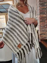 Chal tejido a mano en lana merino color crudo con rayas marrones y flecos en los extremos. Presenta etiqueta de cuero grabada con el logo de la marca.