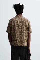 Camisa marrón con estampado floral, corte relaxed fit, cuello bowling, manga corta, bolsillo en el pecho, aberturas laterales y cierre con botones.