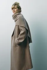 Abrigo largo color beige confeccionado en lana. Presenta cuello solapa con bufanda desmontable, manga larga, bolsillos laterales y cierre frontal cruzado con botones.