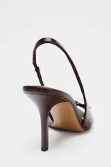 Zapato tipo sandalia destalonada color marrón chocolate, con tacón fino, tira delantera con detalle de hebilla metálica dorada y tira trasera elástica.