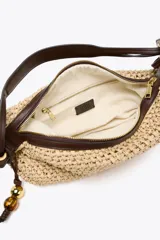 Bolso tipo shopper color beige, con cuerpo trenzado de rafia y asa de hombro de cuero marrón. Tiene un charm colgante en un lateral y cierre con cremallera.