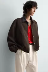 Cazadora de algodón con estructura canvas, color marrón oscuro, de corte relajado. Presenta cuello solapa de cuero negro, cierre frontal con botones a presión, bolsillos de vivo en cadera y puños y bajo elásticos.