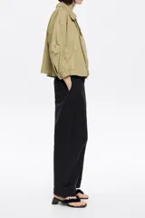 Cazadora corta color verde militar, cruzada con corte fluido, cuello camisero, manga larga recta con botón y cierre frontal con botonadura de presión. Incluye bolsillos plastrón inclinados.