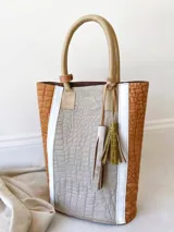 Bolso tipo tote bag de cuero color marrón con textura tipo cocodrilo, detalles en blanco y manijas de cuero color beige. Incluye dos borlas decorativas y correa larga opcional.
