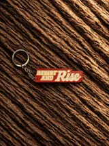 Llavero rectangular con cadena y aro metálico. Diseño con la frase "Resist and Rise" en letras blancas con borde amarillo sobre fondo rojo.