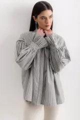 Camisa gris a rayas verticales blancas, de corte amplio, cuello alto con lazo y mangas largas abullonadas con puños elásticos.