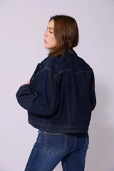 Campera de jean de color azul oscuro, con corte cropped y costuras en contraste color caramelo. Presenta cuello clásico, cierre frontal con botones metálicos y bolsillos a la altura del pecho y laterales.