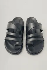 Sandalia plana negra de cuero con diseño de tiras anchas entrecruzadas sobre el empeine y una tira que separa el dedo gordo. Suela plana y gruesa.