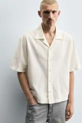 Camisa de denim de algodón, color beige claro, con corte relajado, cuello bowling y manga corta. Presenta cierre frontal con botones a presión y un pequeño bordado a tono en el pecho.