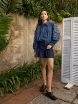 Camisa de denim azul con cuello camisero con volados, mangas largas abullonadas con puños elastizados y bordado blanco en las mangas.