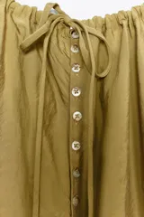 Vestido midi color ocre, de cuello redondo ajustable con cordones y sin mangas. Presenta bolsillos laterales ocultos en la costura y cierre trasero con botones.