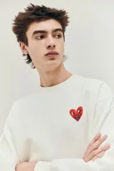 Buzo blanco de corte holgado con cuello redondo y puños y bajo acanalados. Presenta un pequeño logo de corazón rojo con ojos en el pecho.