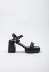 Sandalia de símil cuero negro con taco de 12 cm, cinturilla fina en la parte superior y ajuste con hebilla en el tobillo.