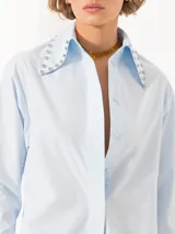 Camisa celeste de poplin, corte clásico, largo a la cadera y mangas largas. Cuello con detalle de piedras engarzadas. Cierre frontal con botones de pasta al tono.
