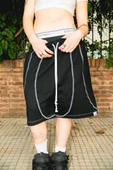 Bermuda de algodón negra, corte baggy, con cordón blanco ajustable en la cintura y costuras blancas a contratono que definen paneles verticales y curvos en los laterales.