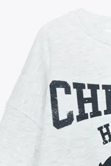 Sudadera gris melange de cuello redondo y manga larga con puños elásticos. Estampado frontal con texto "Chicago Harlem" y dibujo de Betty Boop.