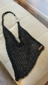 Top tejido a mano en hilo negro con textura de red abierta, escote halter con tiras que se unen a anillos metálicos plateados. Posee una etiqueta de cuero marrón con el nombre de la marca en la base.