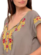 Blusa color taupe de mangas cortas con bordados florales en amarillo y fucsia. Tiene escote redondo con lazo y borlas amarillas.