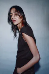 Jersey de punto negro, sin mangas (manga sisa), con cuello cuadrado drapeado y un broche dorado decorativo en el hombro.