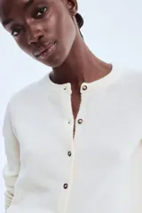 Cárdigan de punto color crema, de manga larga, con cuello redondo y cierre frontal de botones oscuros.