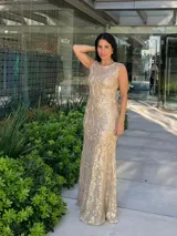Vestido largo sin mangas, color dorado, con base beige y diseño de encaje o brocado con brillo. Corte ajustado al cuerpo que cae recto hasta el suelo.