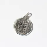 Dije redondo plateado con la imagen de San Benito y texto en latín.