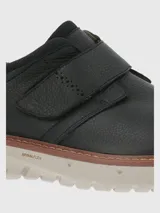 Zapato de cuero negro con ajuste de velcro, punta redonda y suela de goma color crema.