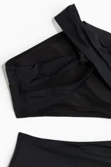 Traje de baño enterizo negro de un hombro con escote asimétrico, bretel ancho, sección cut-out en la cintura, detalle girado en la espalda, copas con relleno extraíble, piernas de corte alto y cobertura media en la parte de atrás.