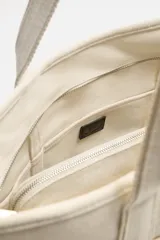 Bolso tipo shopper color crema, fabricado en tejido de algodón. Tiene doble asa corta, compartimento principal con cierre de cremallera, bolsillo interior con cremallera y múltiples bolsillos exteriores.