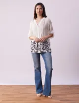 Blusa blanca de gasa bambula con cuello a la base, escote en V y pliegues. Tiene apertura central con botones, mangas ¾ con terminación fruncida y ruedo con detalles bordados en negro.