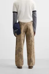 Pantalón de jean color beige con estampado abstracto floral en tonos marrones, de corte amplio, con bolsillos delanteros y traseros, y detalle de tira multifunción en la pierna. Cierre frontal con cremallera y botón.